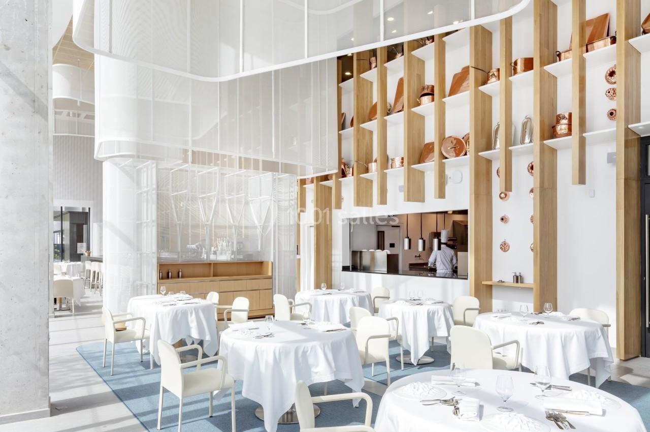 Salle de restaurant lumineuse avec tables dressées, chaises blanches et étagères décorées d'objets en cuivre.