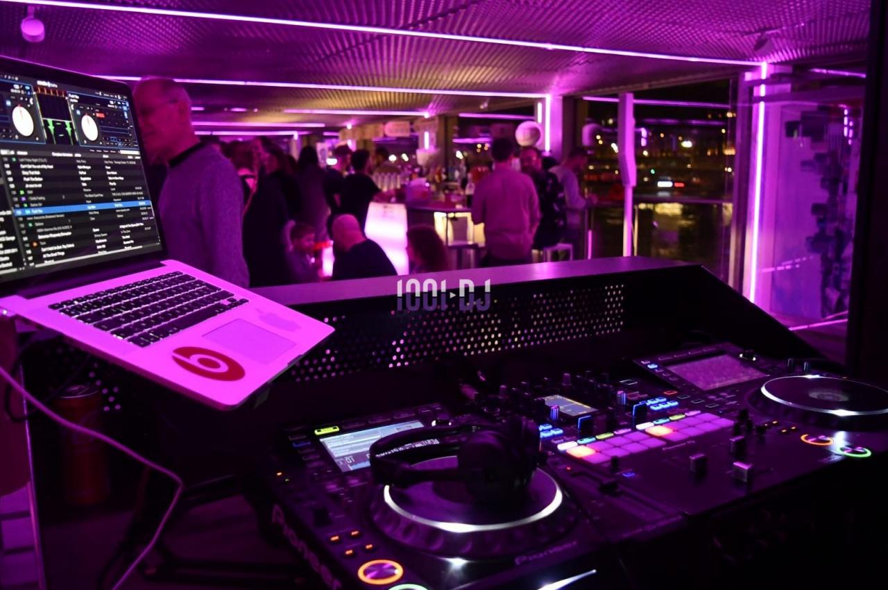 Poste de DJ avec platines et ordinateur dans un espace éclairé par des lumières violettes, avec des personnes en arrière…
