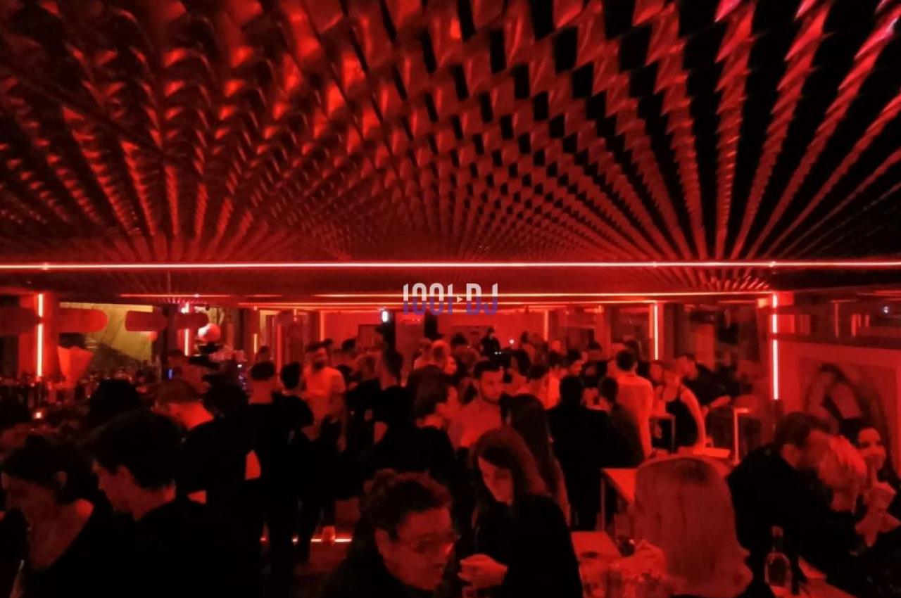 Salle de fête animée avec éclairage rouge, plafond texturé et personnes discutant ou dansant.