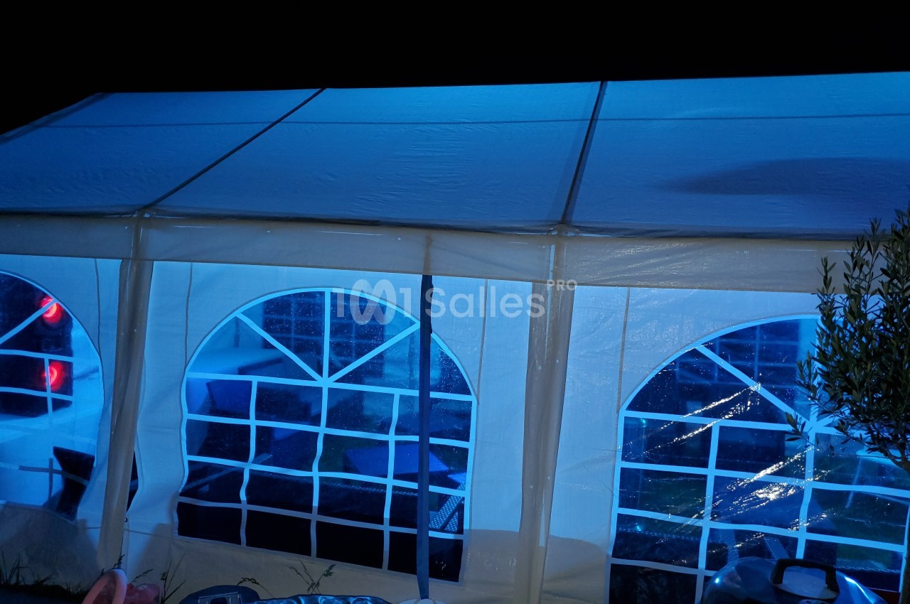 Tente blanche éclairée en bleu la nuit, avec des fenêtres en plastique transparent montrant l'intérieur faiblement visible.