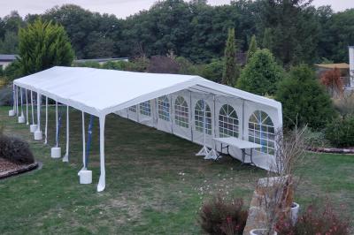 Grande tente blanche rectangulaire installée sur un parking, entourée de végétation et de barrières de sécurité.
