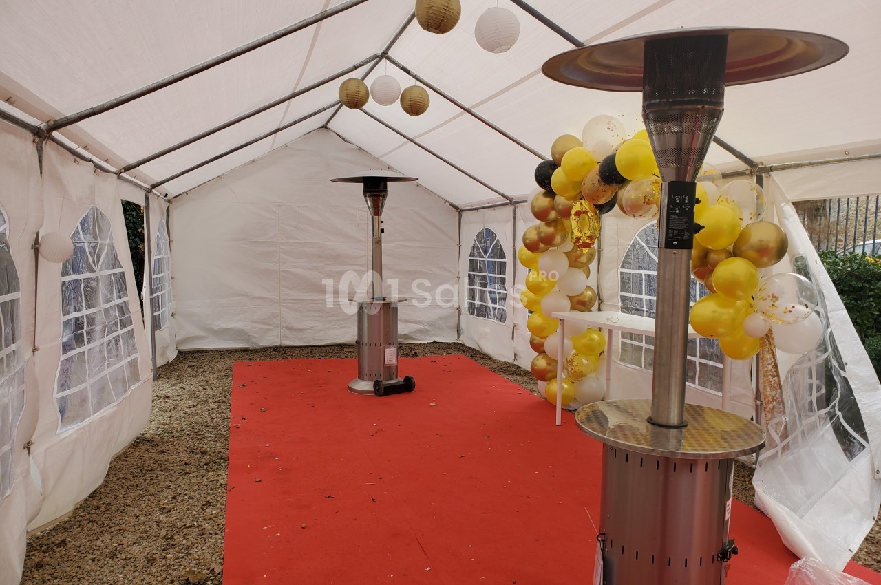 Intérieur d'une tente blanche avec tapis rouge, décorée de ballons jaunes et blancs, et équipée de chauffages.
