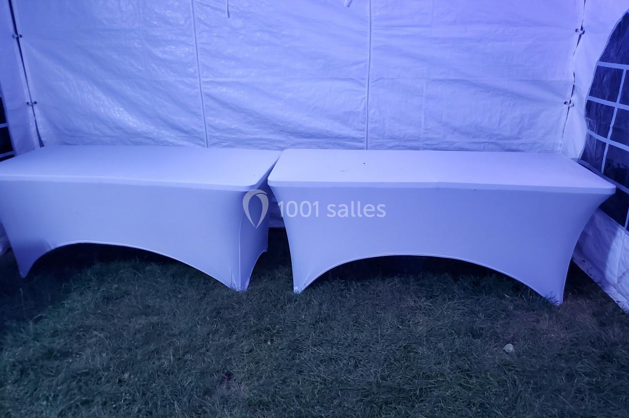 Deux tables rectangulaires recouvertes de housses blanches, disposées sur une pelouse sous une tente blanche.