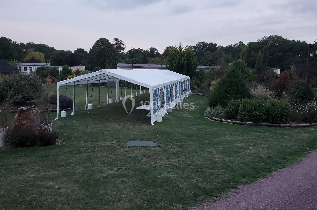 Tente blanche installée sur une pelouse dans un jardin, entourée de végétation et sous un ciel nuageux.