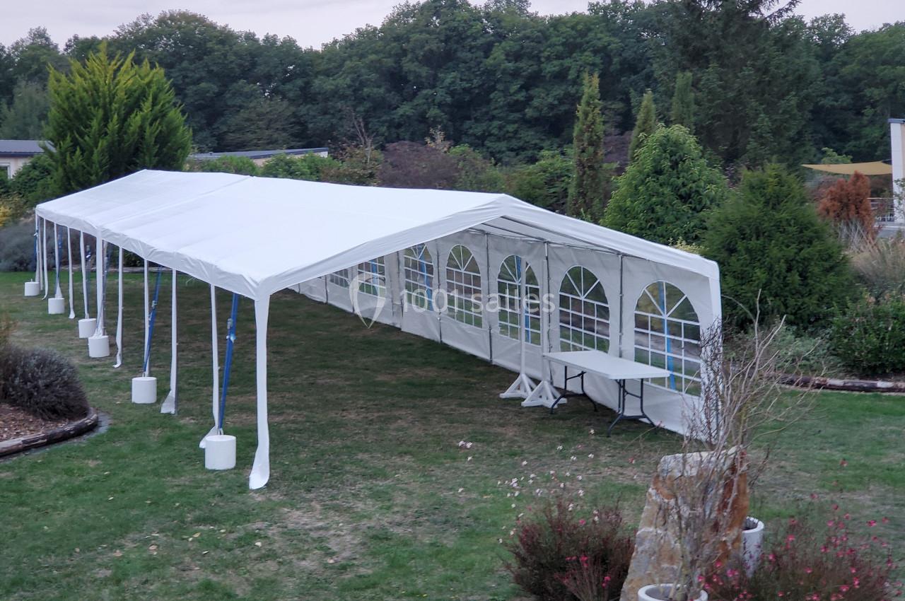 Tente blanche installée dans un jardin, avec des tables visibles à l'intérieur et des arbres en arrière-plan.