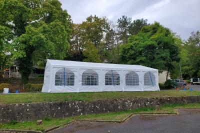 Grande tente blanche avec fenêtres en arc, installée sur un parking entouré de végétation.