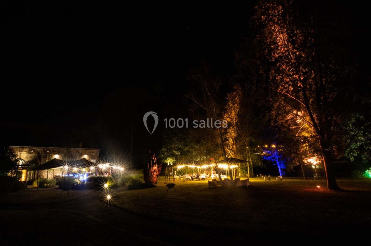 Jardin éclairé la nuit avec guirlandes lumineuses, arbres illuminés et tables disposées en extérieur.