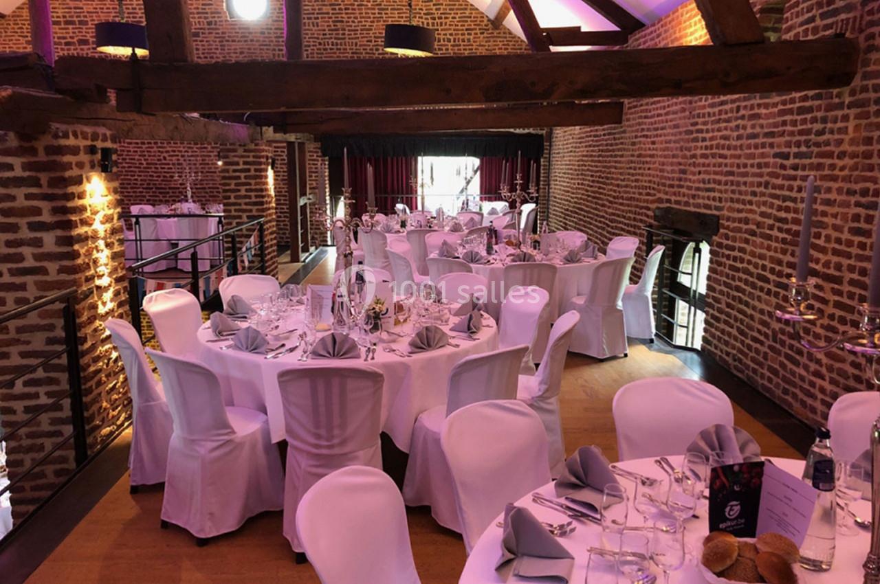 Salle de réception avec murs en briques, tables rondes dressées de nappes blanches et serviettes grises.