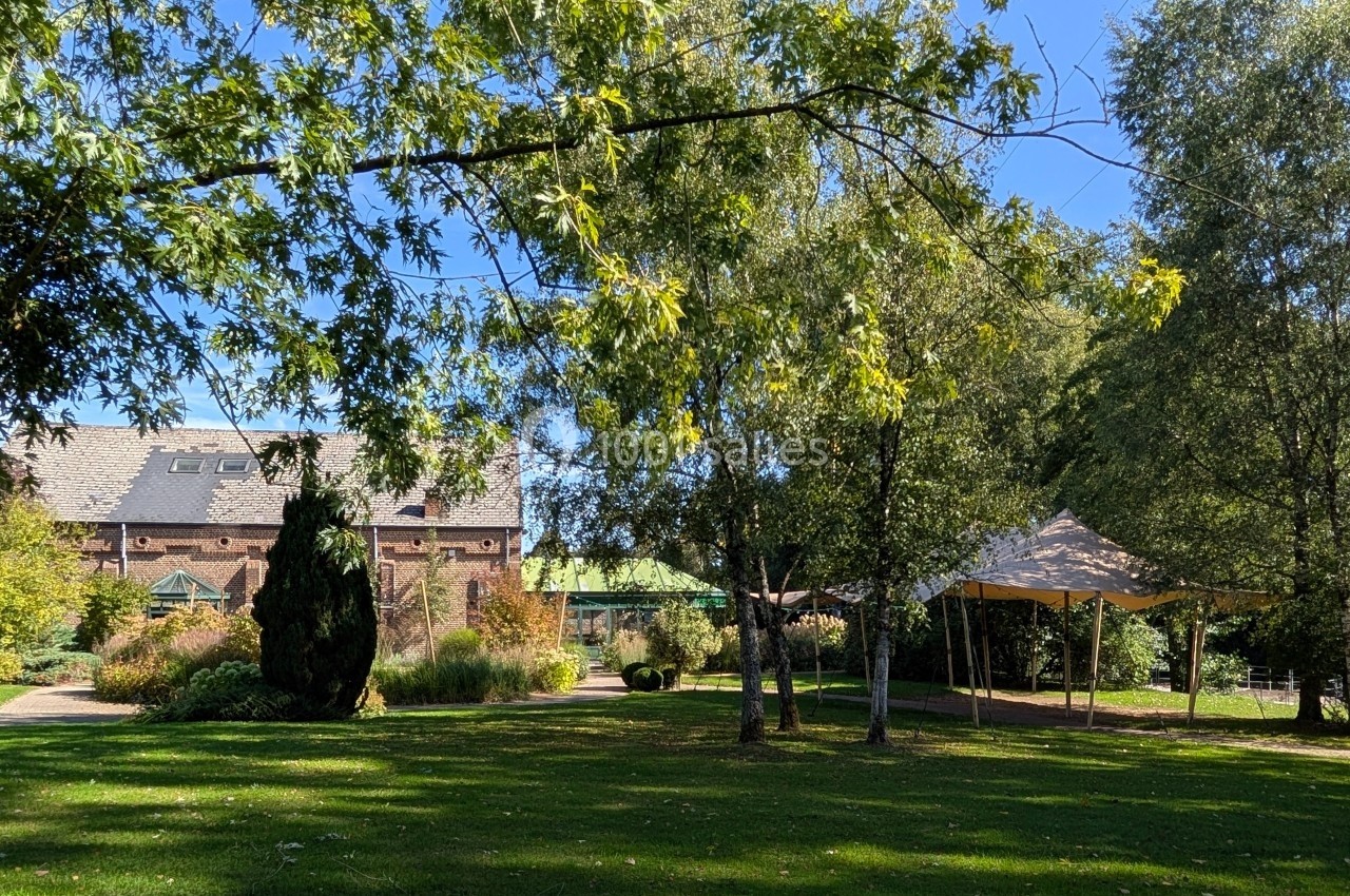 Jardin arboré avec pelouse, arbres, une tente blanche et un bâtiment en briques sous un ciel dégagé.