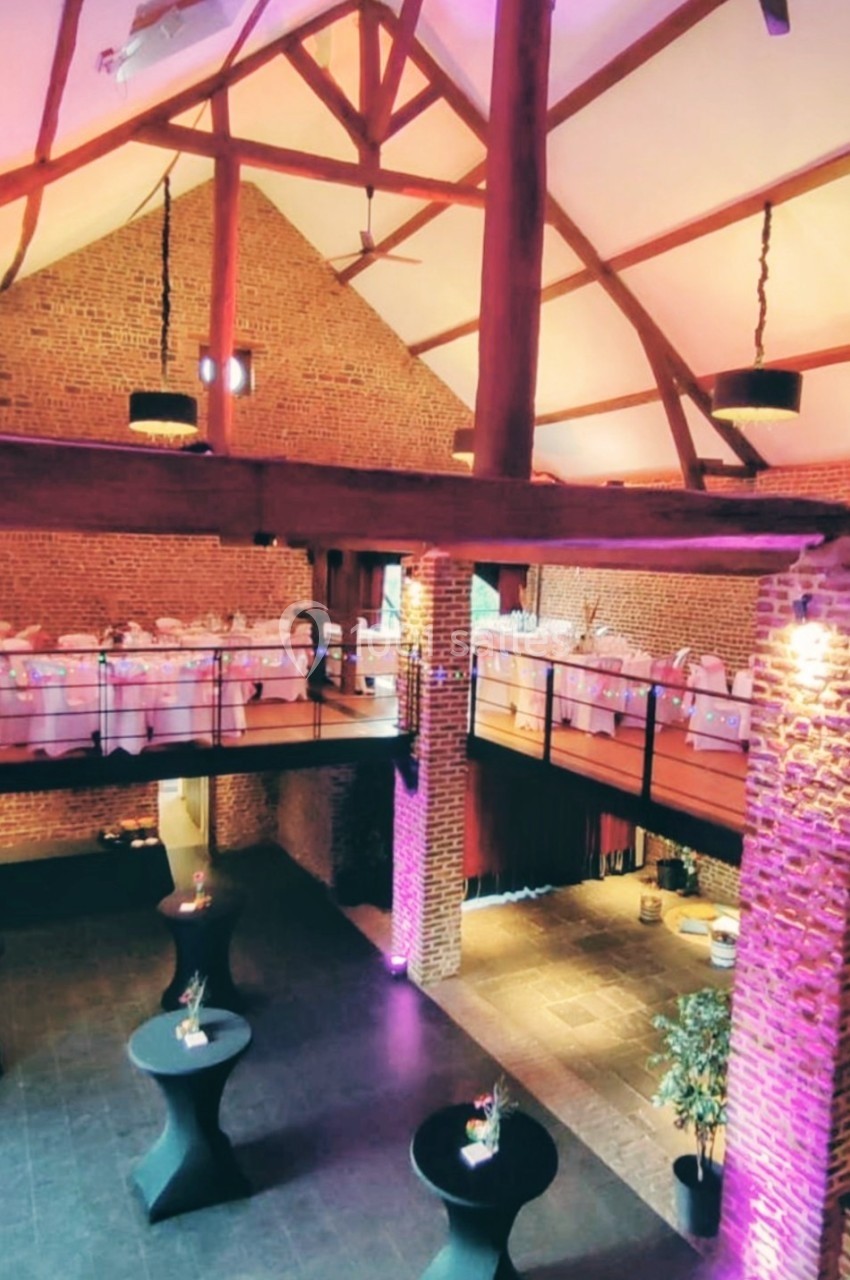 Salle de réception spacieuse avec poutres apparentes, murs en briques, mezzanine et tables hautes décorées.