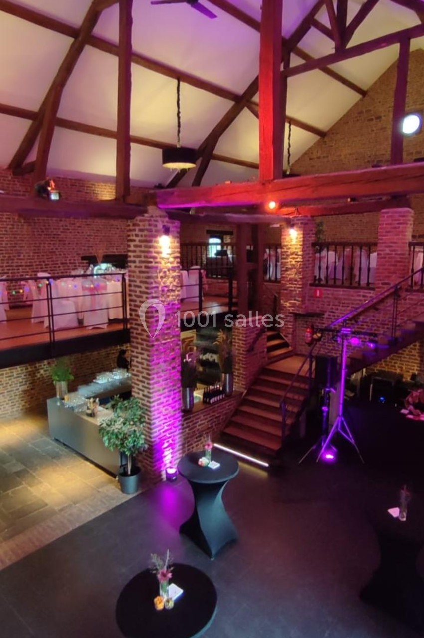 Salle de réception spacieuse avec poutres apparentes, éclairage coloré et tables hautes décorées.