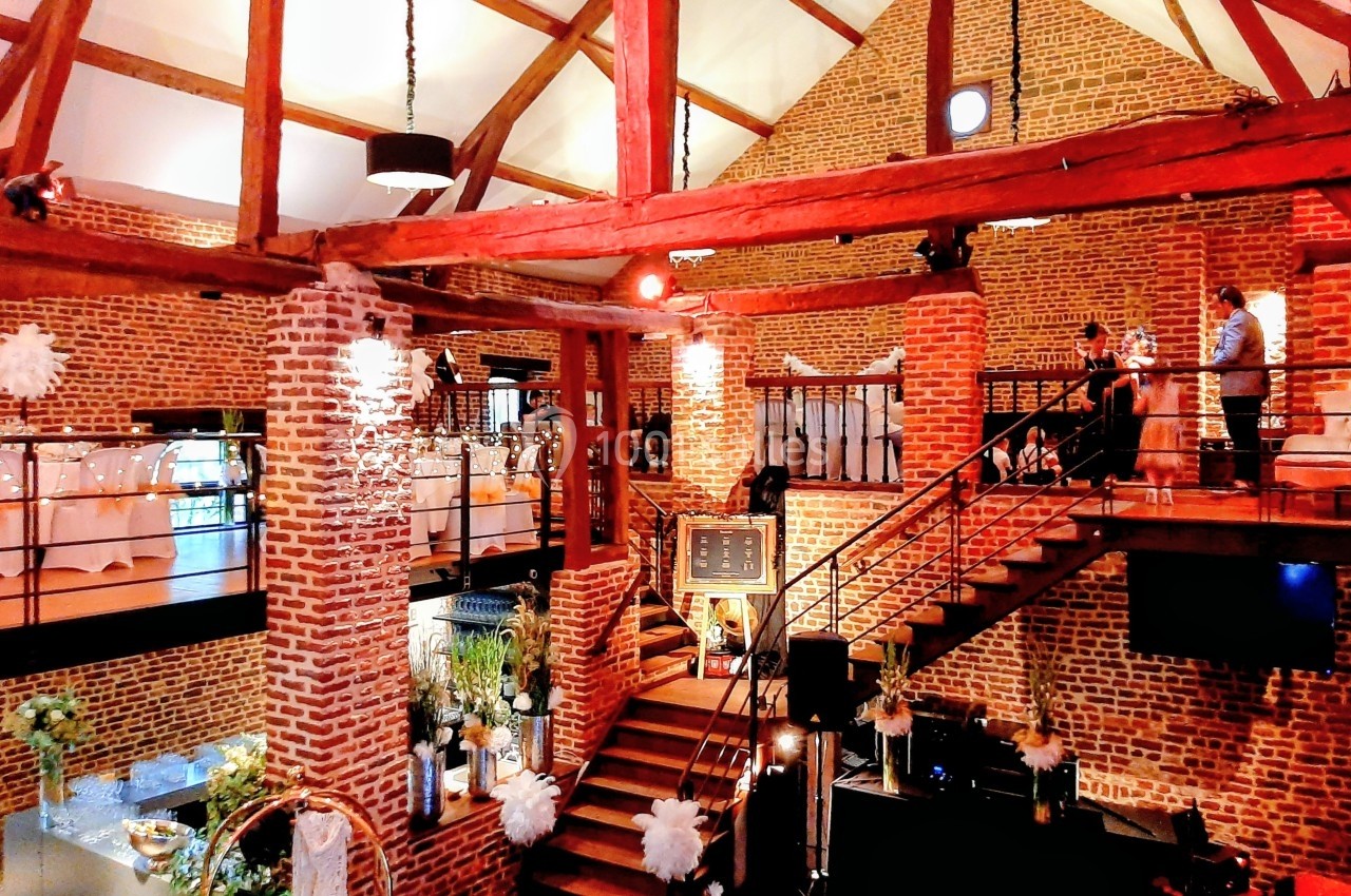 Salle en briques avec poutres apparentes, escalier central en bois, décorations florales et tables dressées pour un…