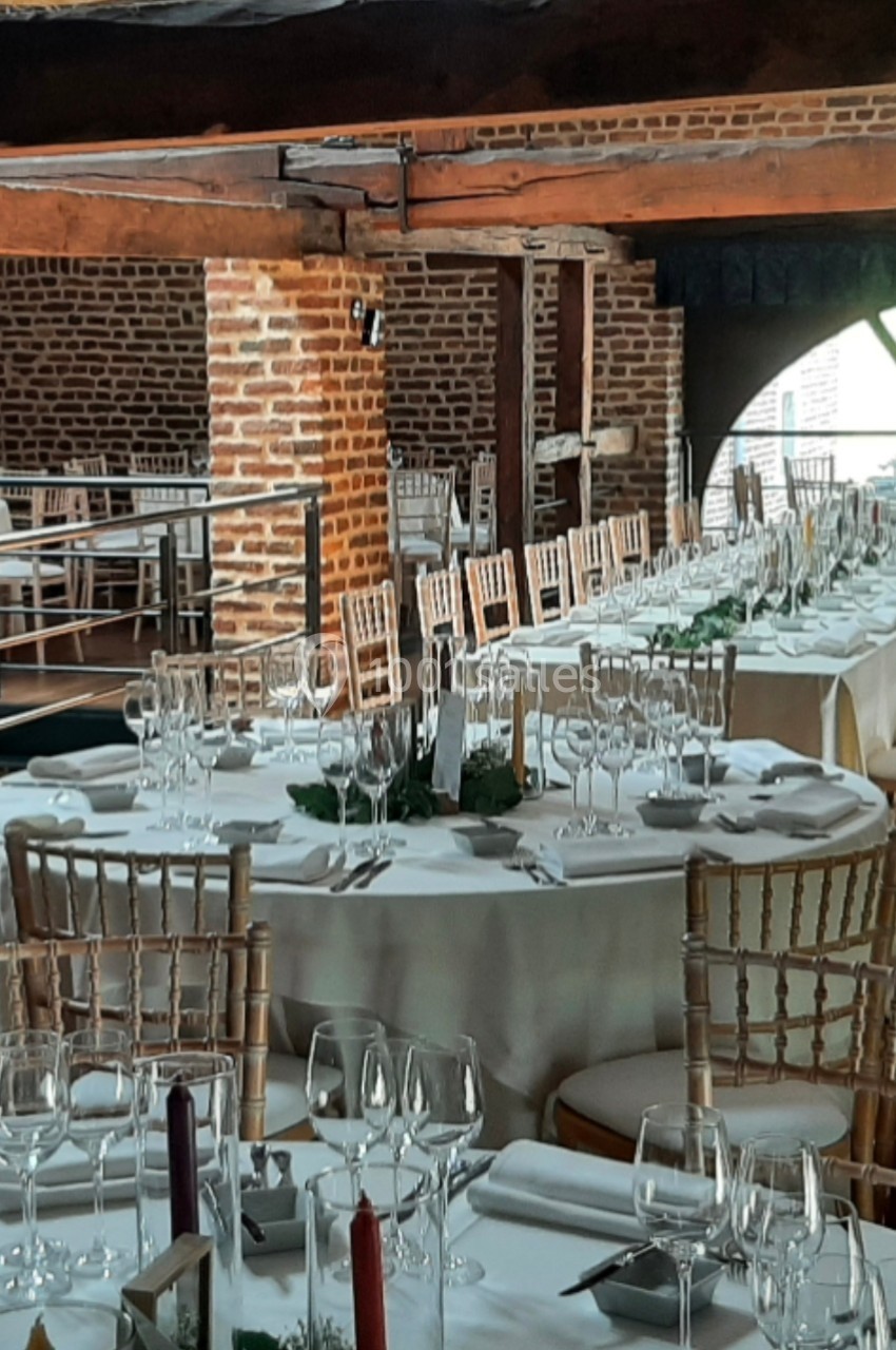 Salle de réception avec tables rondes et rectangulaires dressées, décorée de bougies et de verdure, dans un cadre en briques…