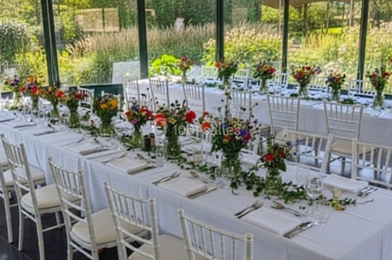 Tables élégamment dressées avec nappes blanches et bouquets de fleurs colorées dans une salle lumineuse entourée de verdure.
