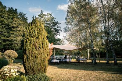 Jardin verdoyant avec arbres, pelouse soignée, tables hautes sous parasols et mobilier coloré près d'un plan d'eau.