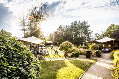 Jardin verdoyant avec arbres, pelouse soignée, tables hautes sous parasols et mobilier coloré près d'un plan d'eau.