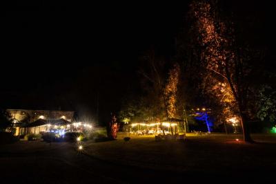 Jardin verdoyant avec arbres, pelouse soignée, tables hautes sous parasols et mobilier coloré près d'un plan d'eau.