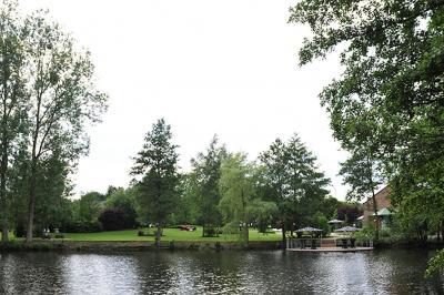 Jardin verdoyant avec arbres, pelouse soignée, tables hautes sous parasols et mobilier coloré près d'un plan d'eau.