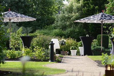 Jardin verdoyant avec arbres, pelouse soignée, tables hautes sous parasols et mobilier coloré près d'un plan d'eau.