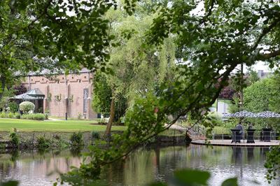 Jardin verdoyant avec arbres, pelouse soignée, tables hautes sous parasols et mobilier coloré près d'un plan d'eau.
