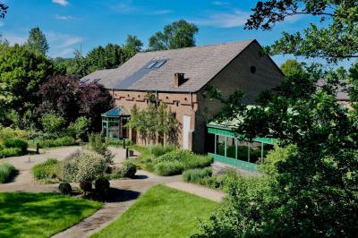 Jardin verdoyant avec arbres, pelouse soignée, tables hautes sous parasols et mobilier coloré près d'un plan d'eau.