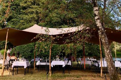 Jardin verdoyant avec arbres, pelouse soignée, tables hautes sous parasols et mobilier coloré près d'un plan d'eau.