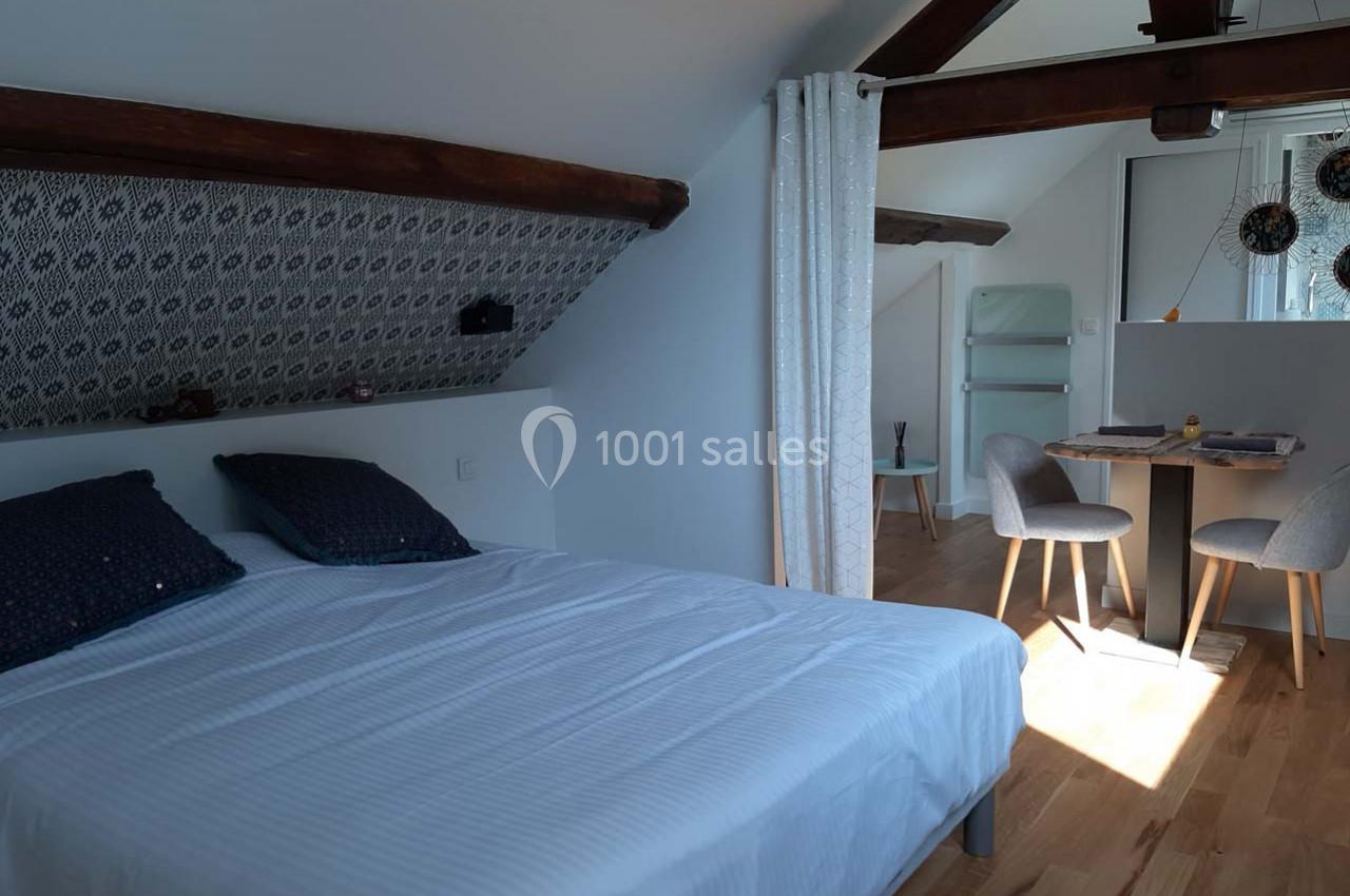 Chambre mansardée avec lit double, table à manger, chaises et poutres apparentes, éclairée par une fenêtre.