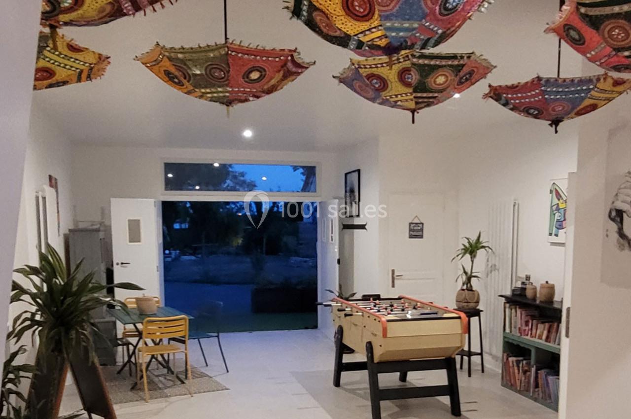 Salle lumineuse avec baby-foot, plantes, étagères de livres et parapluies colorés suspendus au plafond.
