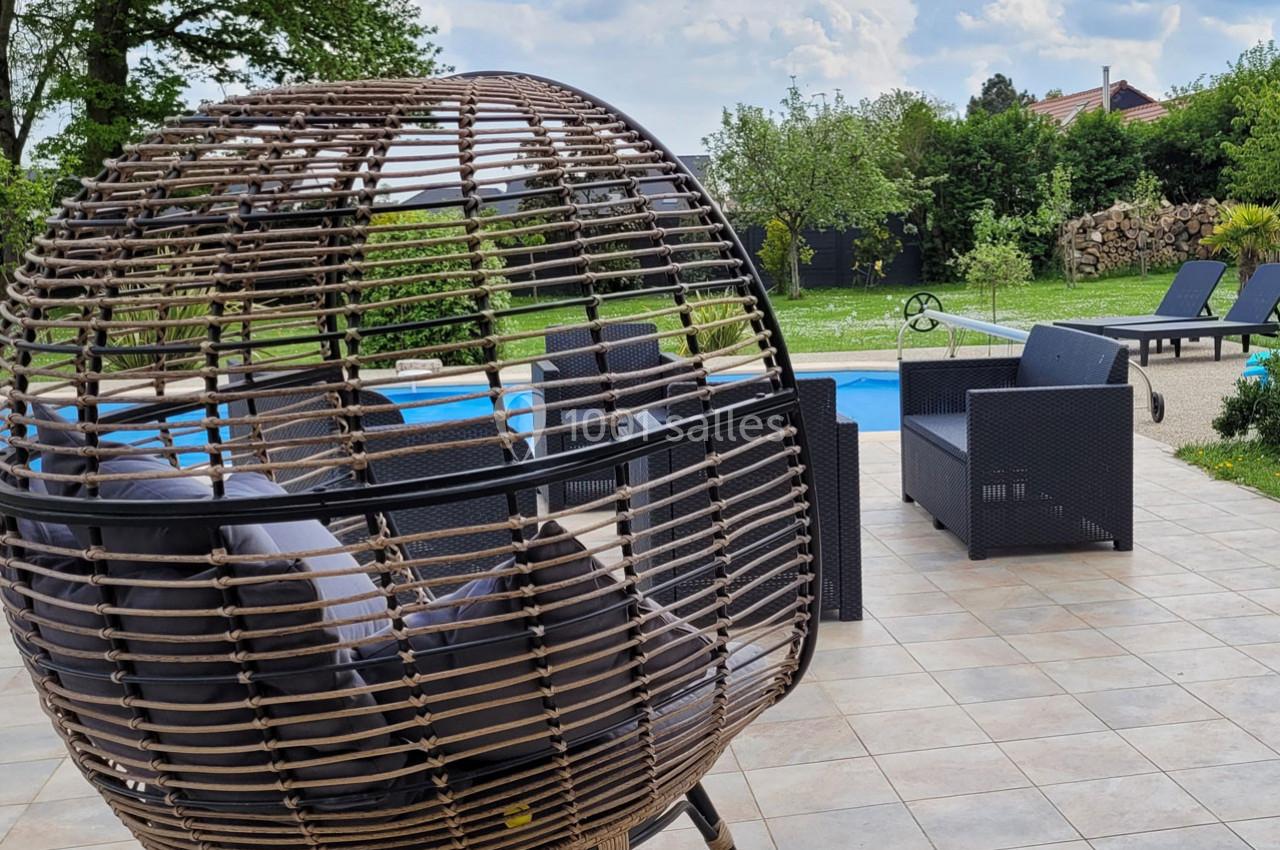 Fauteuil suspendu en rotin sur une terrasse carrelée, avec piscine, salon de jardin et espace vert en arrière-plan.