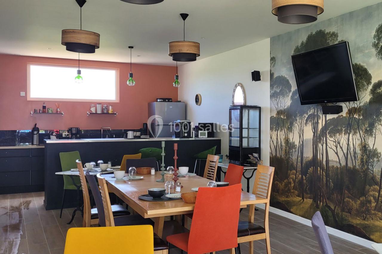 Salle à manger moderne avec table dressée, chaises colorées, cuisine ouverte et mur décoré d'une fresque forestière.