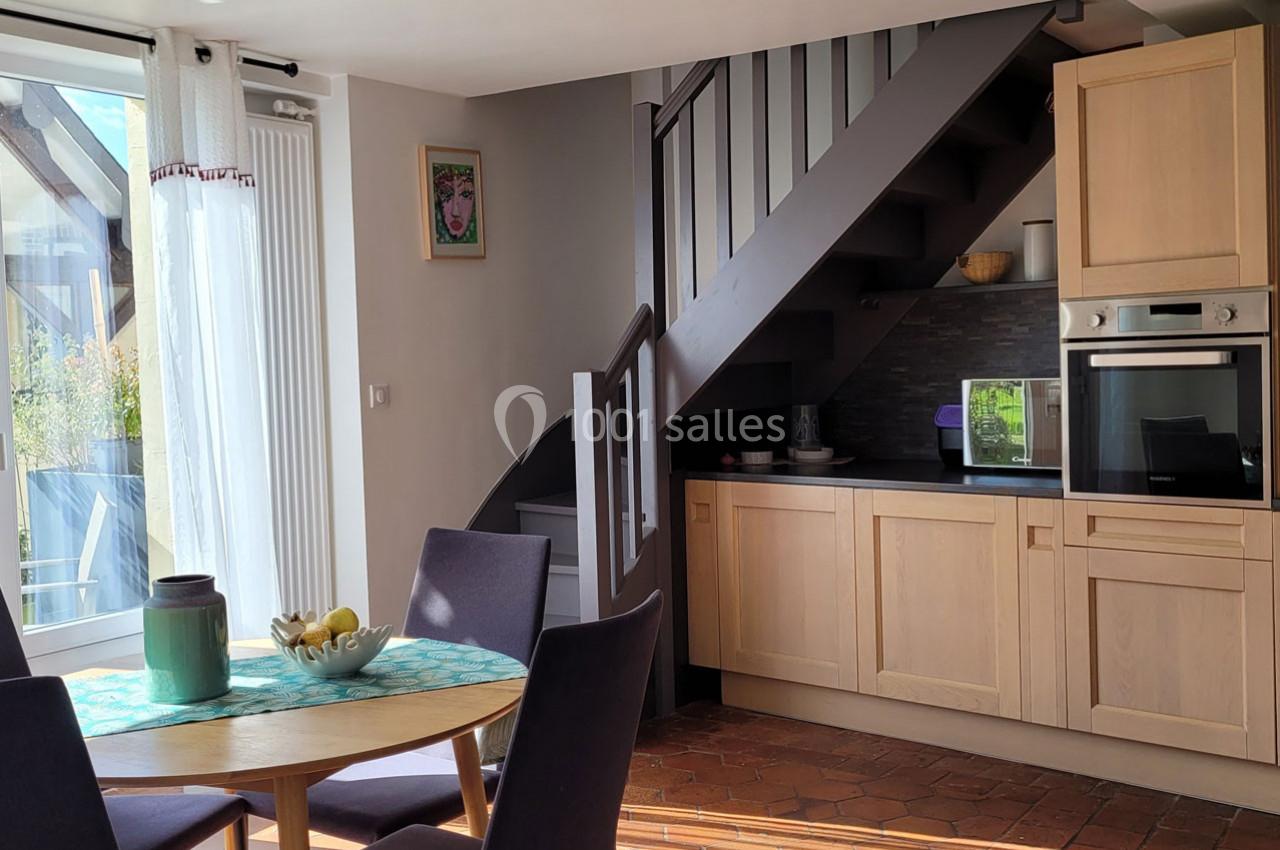 Cuisine ouverte avec escalier en bois, table ronde dressée et lumière naturelle entrant par une baie vitrée.