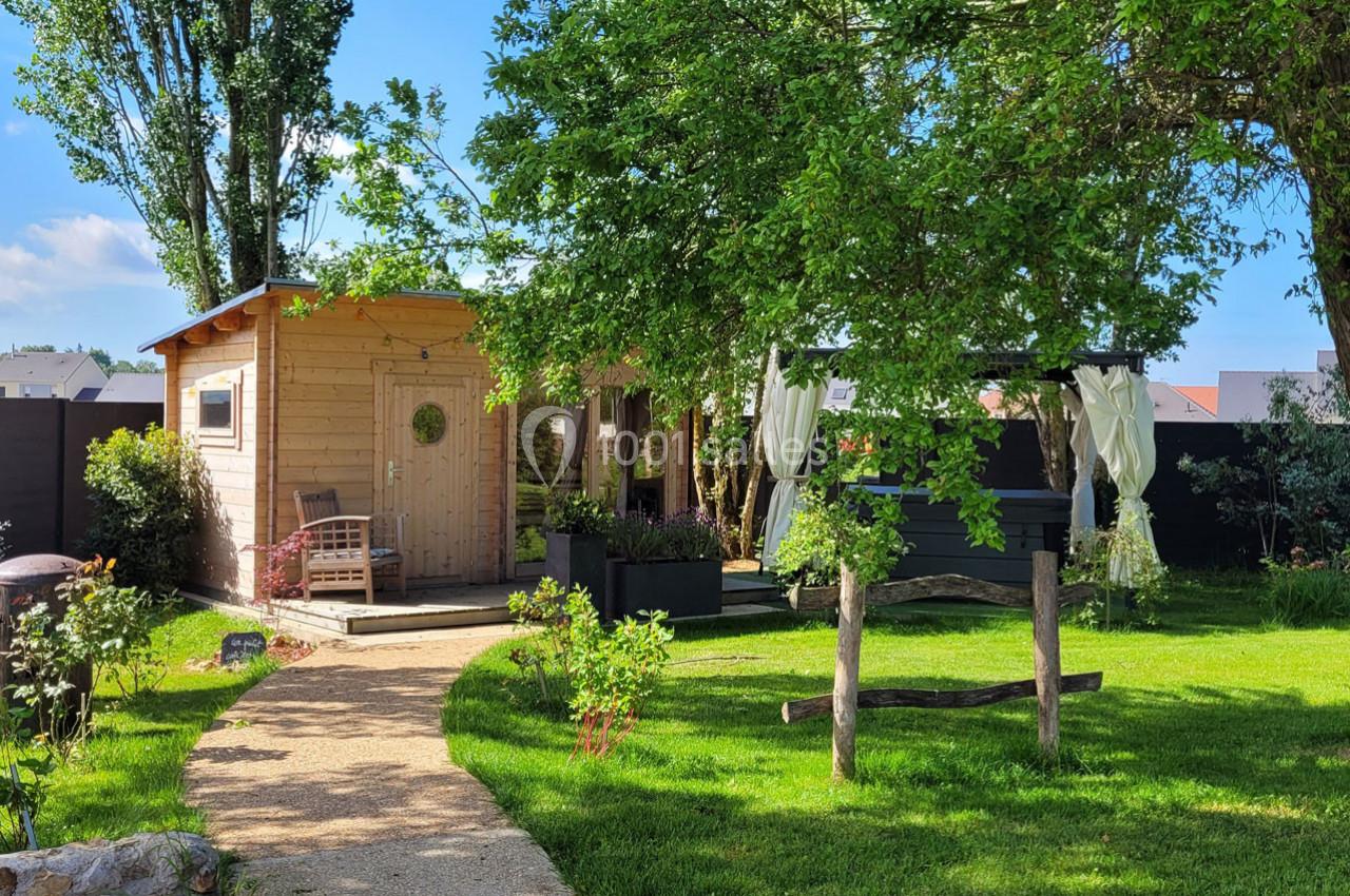 Abri en bois et pergola avec rideaux blancs dans un jardin verdoyant, chemin en pierre menant à l'entrée.