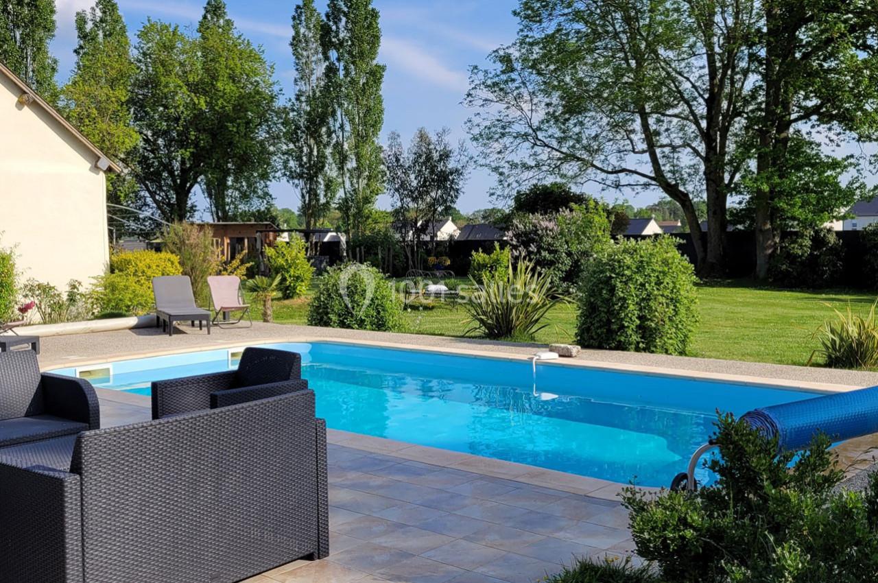 Piscine extérieure entourée de mobilier de jardin, pelouse et arbres sous un ciel dégagé.
