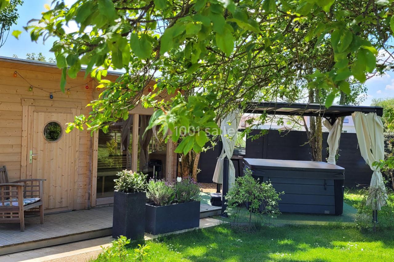 Cabane en bois avec terrasse, entourée de verdure, un spa et un espace ombragé avec rideaux dans un jardin.