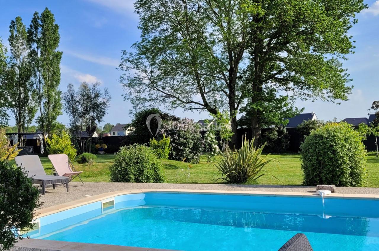 Piscine extérieure entourée de verdure avec des chaises longues, un jardin arboré et un ciel dégagé en arrière-plan.