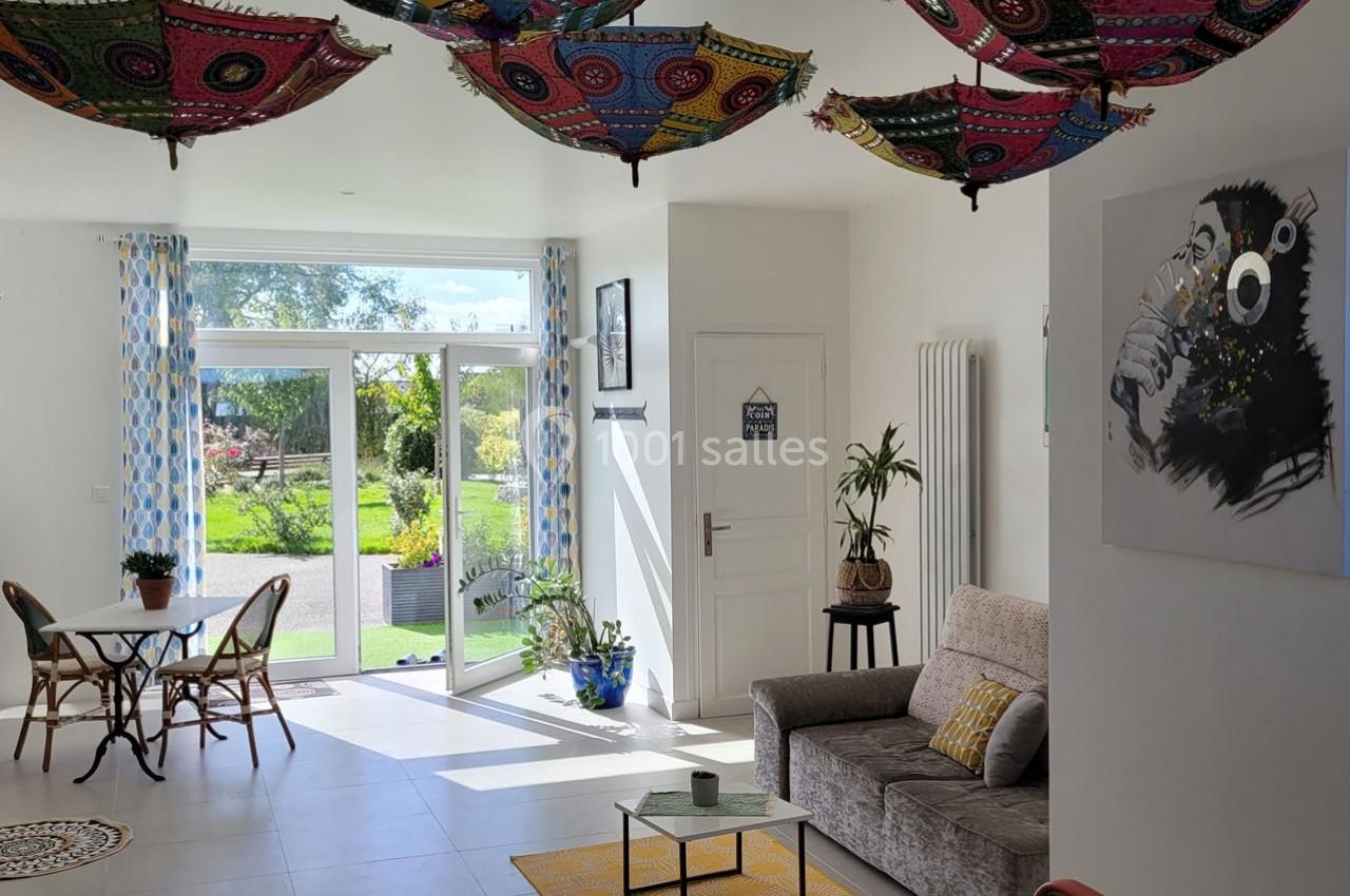 Salon lumineux avec canapés, table ronde, plantes, grandes baies vitrées et parasols colorés suspendus au plafond.