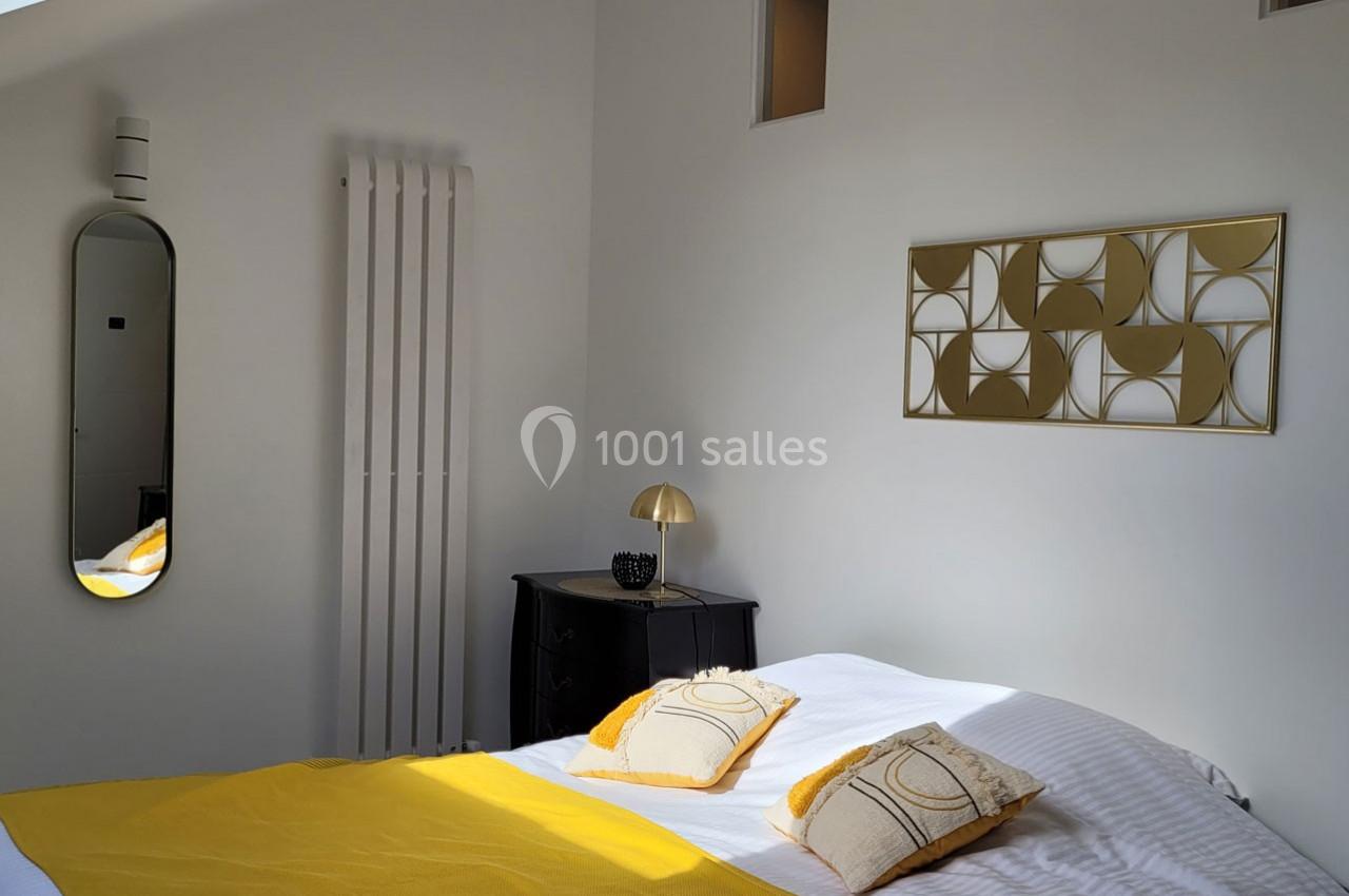 Chambre lumineuse avec lit double, couvre-lit jaune, coussins assortis, miroir, radiateur et décoration murale dorée.
