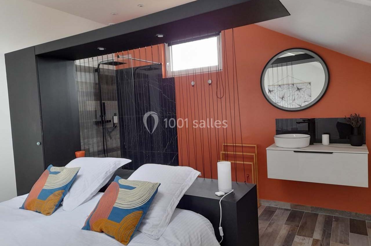 Chambre moderne avec lit double, cloison ajourée, mur orange, salle de bain attenante et lavabo sur meuble suspendu.