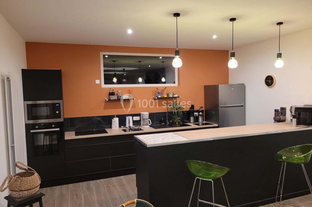 Cuisine moderne avec mur orange, meubles noirs, plan de travail clair, électroménagers en inox et éclairage suspendu.