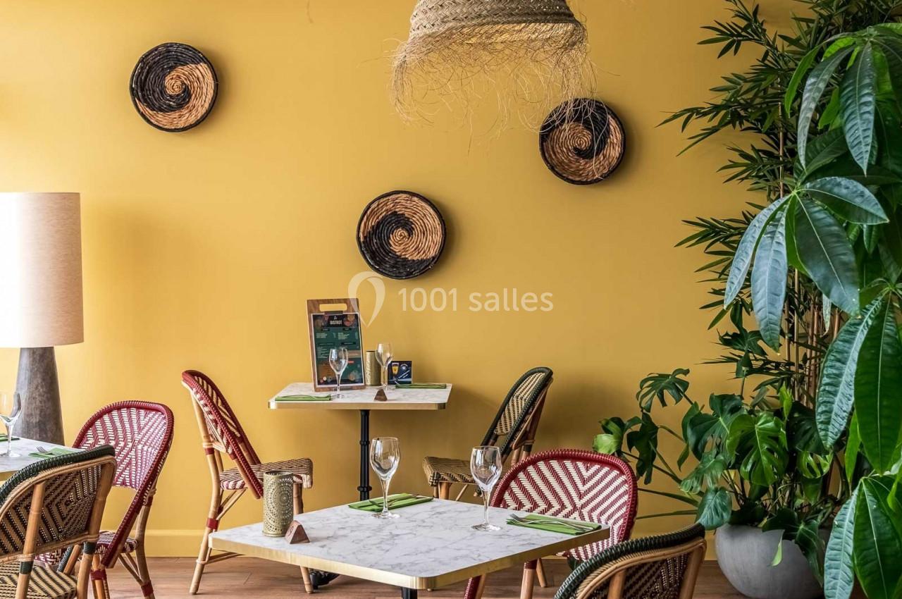 Salle de restaurant avec murs jaunes, tables en marbre, chaises en rotin, plantes vertes et décorations murales tressées.