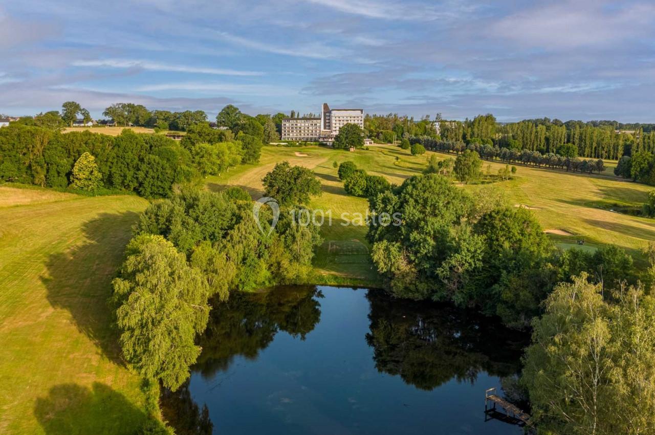 Vue aérienne d'un paysage verdoyant avec un étang au premier plan et un grand bâtiment au loin entouré d'arbres.