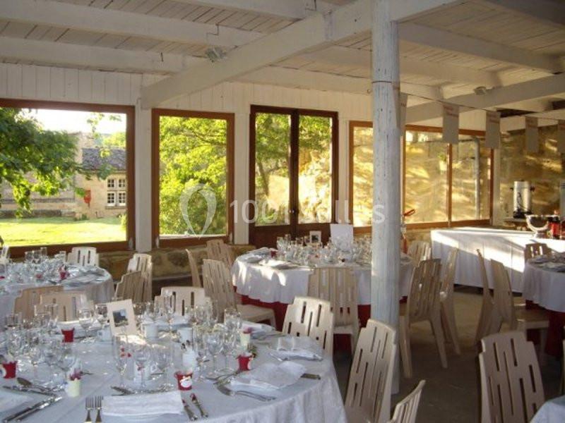 Salle lumineuse décorée pour un repas, avec tables rondes dressées et grandes fenêtres donnant sur un jardin.