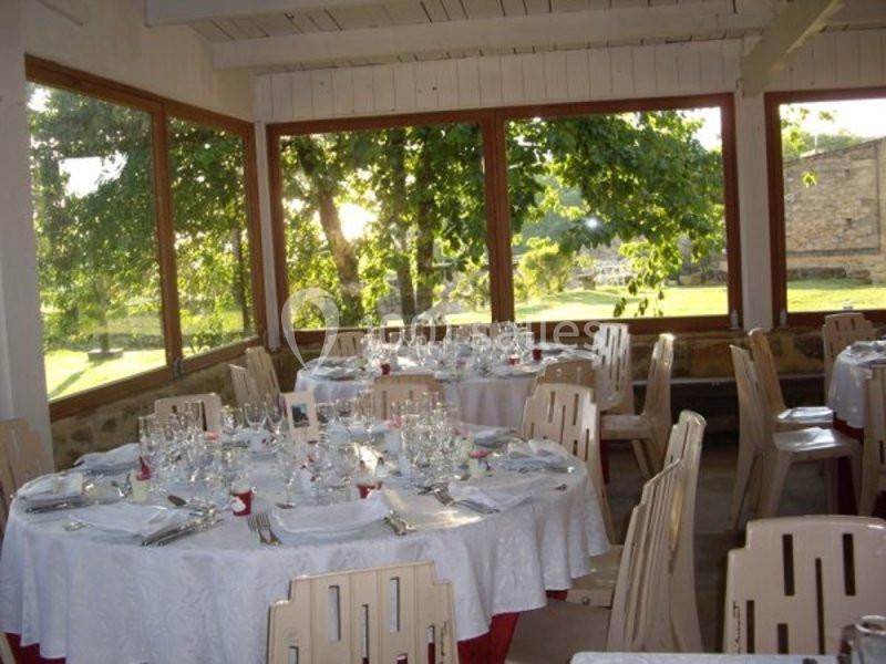 Salle lumineuse avec grandes baies vitrées, tables rondes dressées avec nappes blanches et chaises en bois clair.