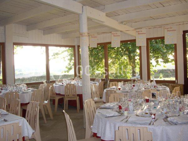 Salle lumineuse avec grandes baies vitrées, tables rondes dressées avec nappes blanches et chaises en bois clair.