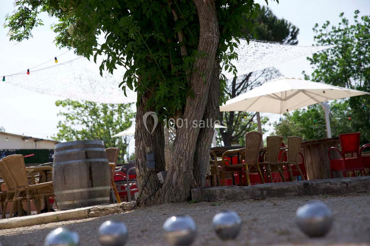 Terrain de pétanque ombragé avec des boules au sol, entouré de chaises, tables et parasols en arrière-plan.