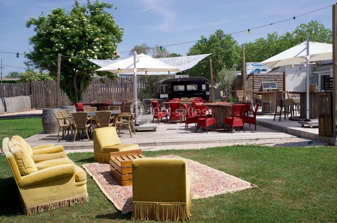 Espace extérieur aménagé avec tables, chaises, fauteuils et parasols, entouré de verdure et éclairé par des guirlandes.