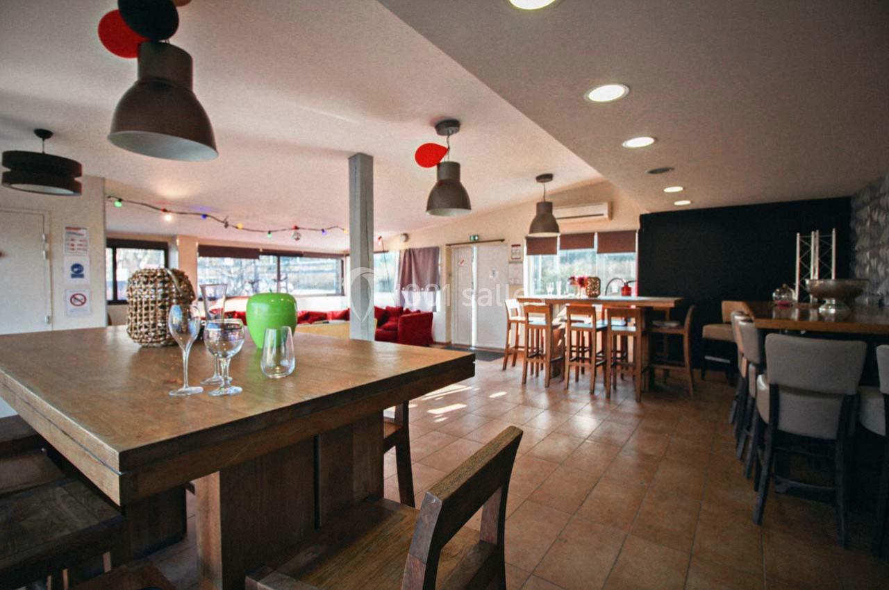 Salle lumineuse avec tables en bois, chaises, suspensions modernes et baies vitrées donnant sur l'extérieur.