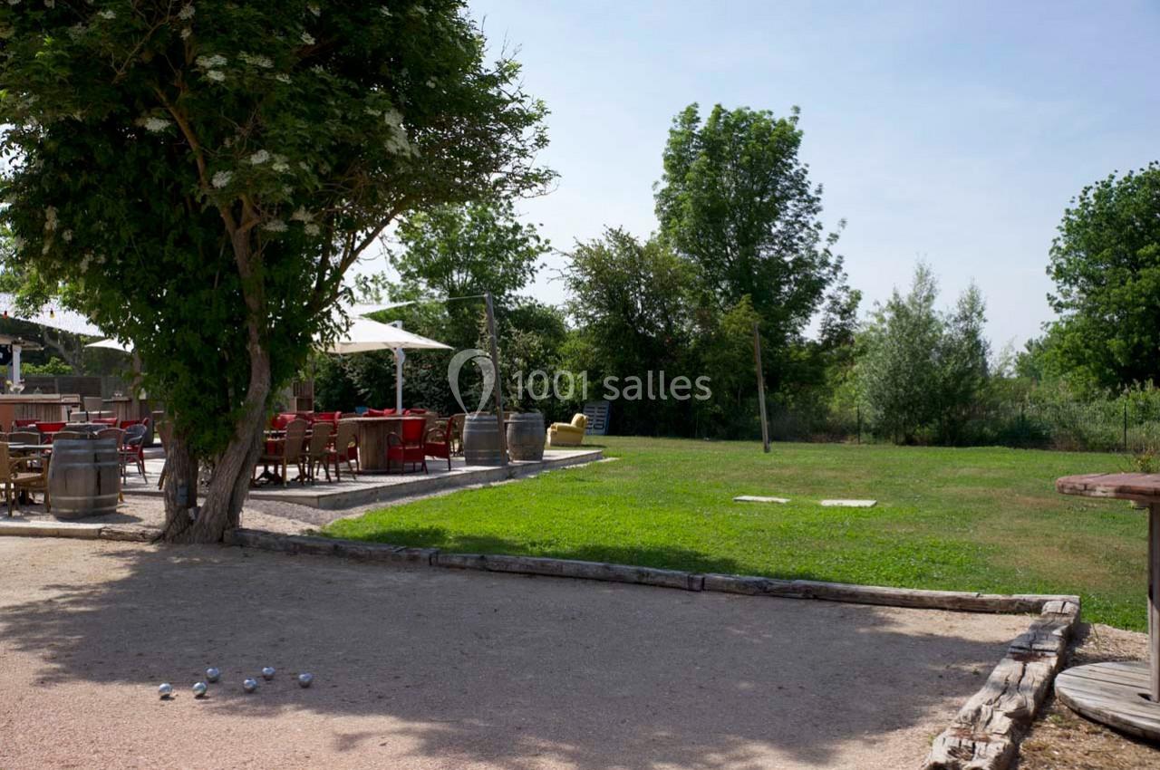 Terrain de pétanque ombragé avec des boules au sol, à côté d'une terrasse extérieure aménagée et d'une pelouse.