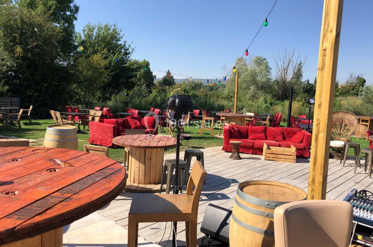 Terrasse extérieure avec tables en bois, tonneaux, canapés rouges et guirlandes lumineuses, entourée de verdure.