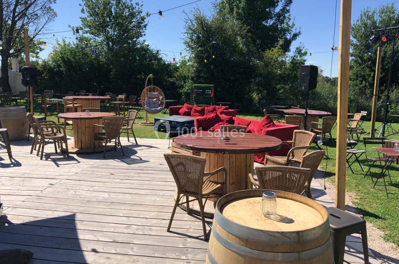 Terrasse en bois avec tables, chaises en rotin, tonneaux et fauteuils rouges, entourée de verdure et guirlandes lumineuses.